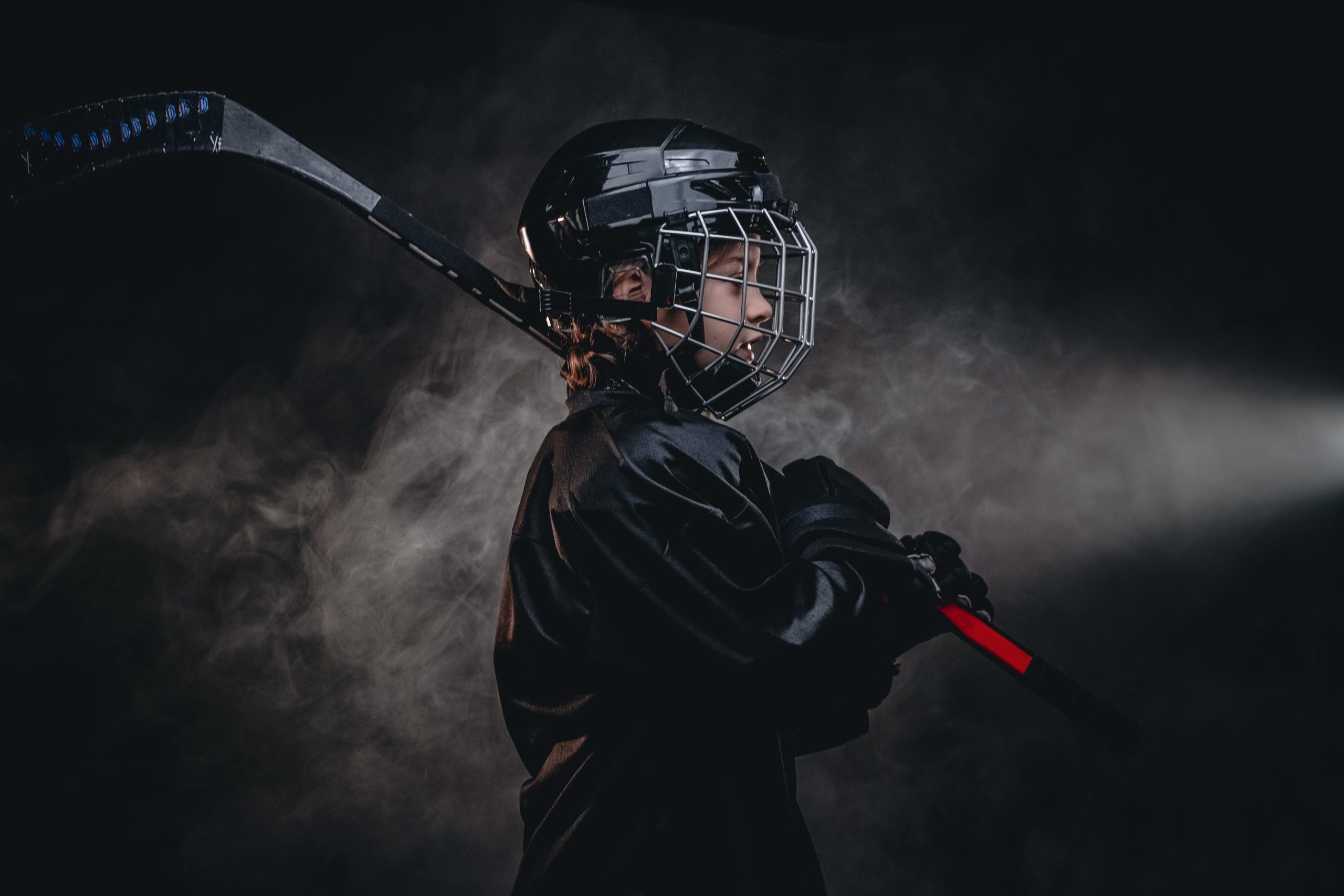 Young blonde sporty boy, ice hockey player, posing in a dark studio for a photoshoot, wearing an ice-skating uniform, helmet, gloves and holding up a hockey stick, confident look from the side